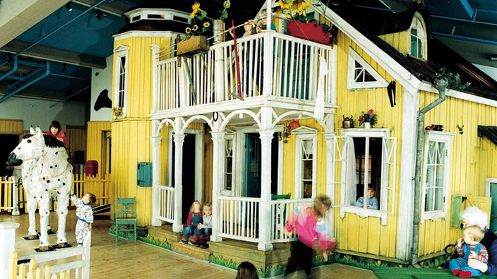 Kinder spielen vor und in einem gelben Holzhaus, das in einer Halle steht. Links vor dem Haus, das die Villa Kunterbunt darstellen soll, steht ein weißes Holzpferd, auf dem ein Kind reitet.