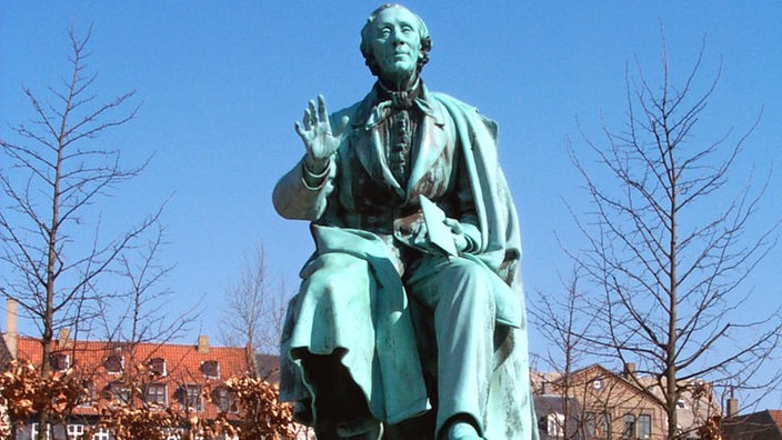 Eine Statue des Dichters Hans Christian Andersen auf einem Sockel. Die Figur sitzt und hat ihren rechten Arm erhoben.