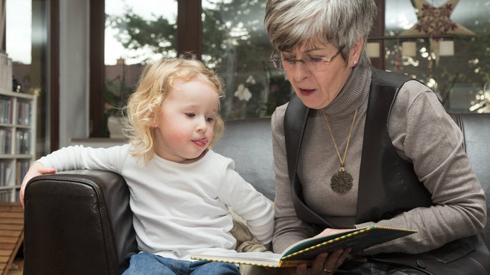 Symbolbild: Eine ältere Frau liest ihrem Enkelkind aus einem Buch vor. 