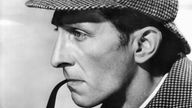 Der britische Schauspieler Peter Cushing auf einem Schwarzweiß-Photo. Das Brustbild zeigt ihn in der Filmrolle des Sherlock Holmes mit einer karierten Tweedmütze und einem karierten Regencape. Seine linke Hand hält eine Pfeife an den Mund.