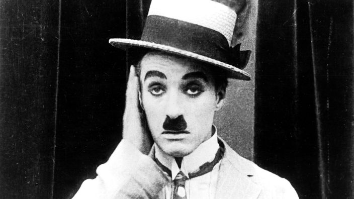 Charlie Chaplin hält sich ein Ohr zu.