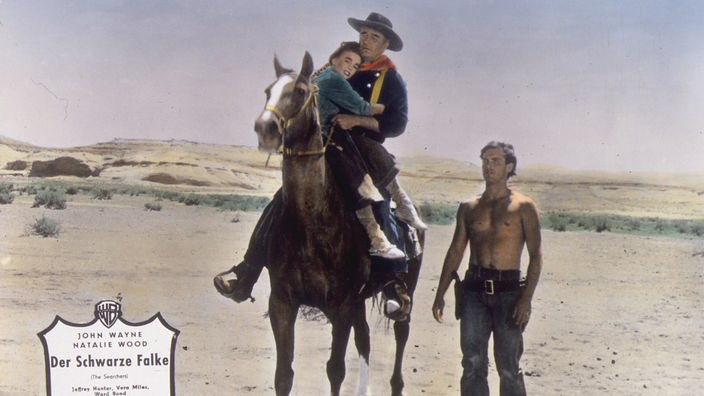 Ein Cowboy reitet auf einem Pferd und hält eine Frau im Arm. Daneben geht ein Mann mit freiem Oberkörper.