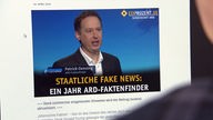 Eine Person schaut auf einen Monitor, auf dem 'Staatliche Fake News: Ein Jahr ARD-Faktenfinder' unter dem Bild eines Mannes steht.