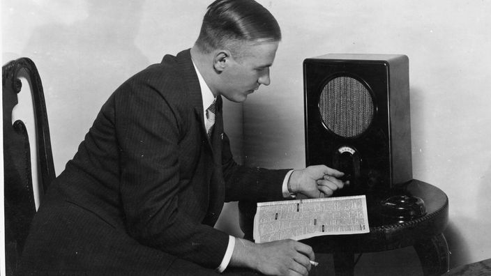 Ein Mann sitzt vor einem Radiogerät, an der Wand hängen Fotos von Hitler und Goebbels.