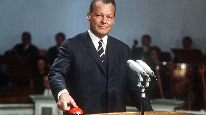 Willy Brandt steht in einem blauen Anzug vor einem Pult. Er drückt auf einen roten Knopf, der symbolische Auslöser für den Start des Farbfernsehens.