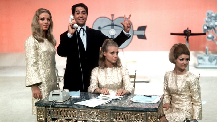 Vico Torriani im Anzug mit Telefonhörer in der Hand, um ihm herum sitzen seine Assistentinnen um einen Tisch