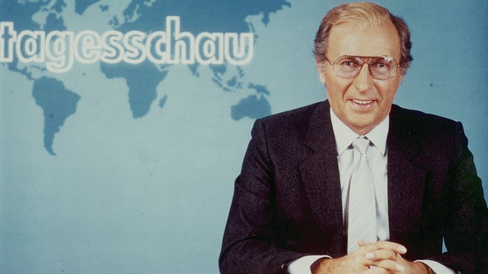 ARD-Nachrichtensprecher Karl Heinz Köpcke sitzt in einem schwarzen Anzug im Tagesschau-Studio (1984).