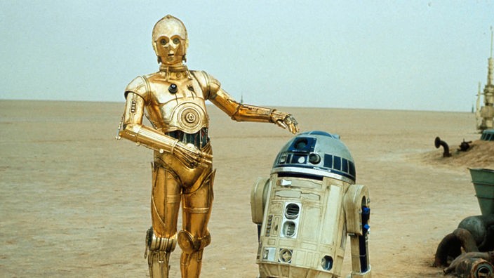 Szene aus dem Film 'Krieg der Sterne'. Die beiden Roboter R2D2 und C-3PO stehen nebeneinander in einer Wüstenlandschaft.