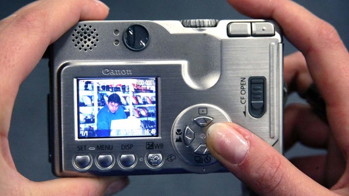 Frauenfinger halten eine Digitalkamera. Der Betrachter kann auf dem Display verschwommen ein Foto sehen.