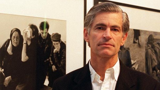 Der Fotograf James Nachtwey schaut direkt in die Kamera. Im Hibtergrund hängen einige seiner Bilder in einer Ausstellung. | Bildquelle: dpa Der Fotograf James Nachtwey schaut direkt in die Kamera. Im Hibtergrund hängen einige seiner Bilder in einer Ausstellung.