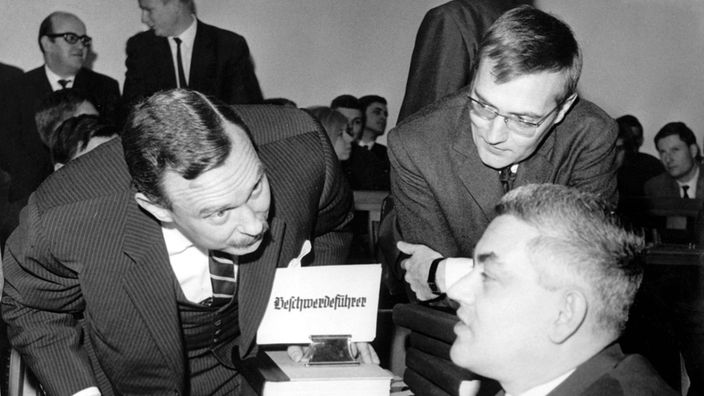 Schwarzweiß-Foto: Drei Männer im Anzug unterhalten sich. Zwischen ihnen steht ein Tisch, auf dem viele Akten liegen. Außerdem steht auf dem Tisch ein Schild, auf dem in Frakturschrift 'Beschwerdeführer' steht.