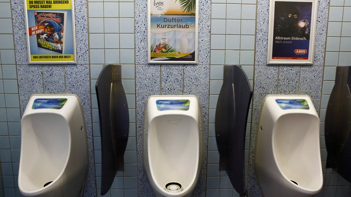 Vor Pissoirs in einer Herrentoilette hängen Werbeplakate