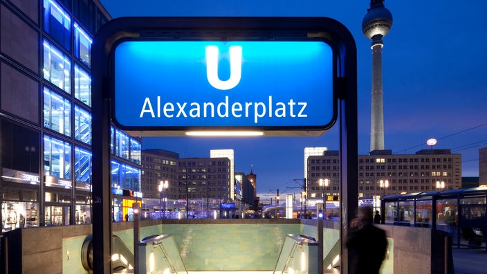 U-Bahnhaltestellenschild "Alexanderplatz". Dahinter der Eingang zur U-Bahn und im Hintergrund der Berliner Fernsehturm.