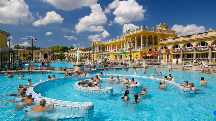 Im Hintergrund ist der einem Schloss ähnelnde gelbe Bau des Széchenyi-Bads zu sehen, im Vordergrund baden die Menschen in Wasseranlagen, die aussehen wie Barockgärten.