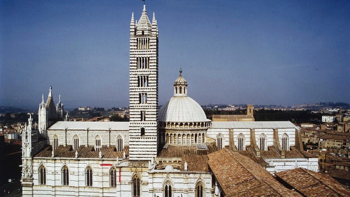 Blick von schräg oben auf den Dom von Siena. In der Mitte des Doms ist eine hellgraue Kuppel zu sehen, direkt daneben der schwarzweiß gestreifte Glockenturm.