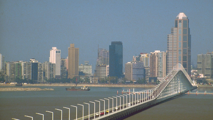 Blick über Wasser auf die von Skyline von Macau. Im Vordergrund führt eine große Brücke in die Stadt.