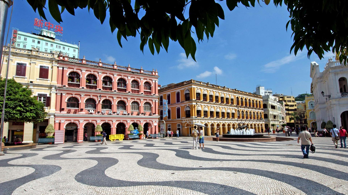 Blick auf einen Platz, der durch prächtige, bunte Kolonialbauten begrenzt wird. Der Platz ist in Wellen schwarzweiß gepflastert. | Bildquelle: Macau-Info Blick auf einen Platz, der durch prächtige, bunte Kolonialbauten begrenzt wird. Der Platz ist in Wellen schwarzweiß gepflastert.