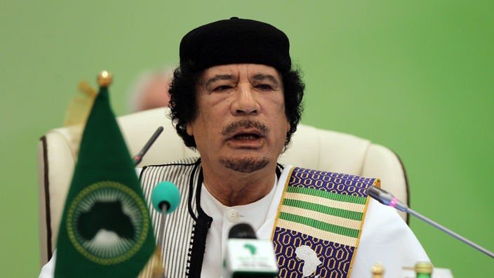 Muammar al-Gaddafi – weißer Kaftan, bunte Schärpe, schwarze Kappe – vor Mikrofonen und einer Flagge der Afrikanischen Union