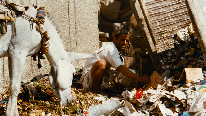 Mann hockt auf der Straße und wühlt in Müll. Links von ihm steht ein weißer Esel.