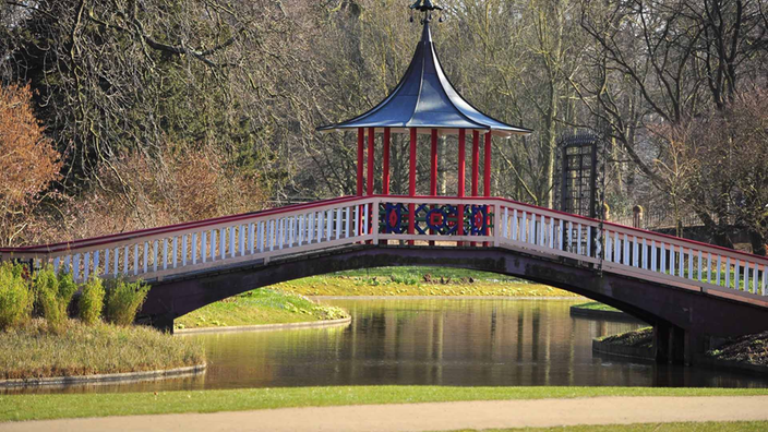 Eine zierliche gebogene Fußgängerbrücke in den Farben Weiß, Rot und Blau über einem ruhigen Gewässer im Park. Die Brückenmitte krönt ein kleiner offener Pavillon mit spitzem Dach.