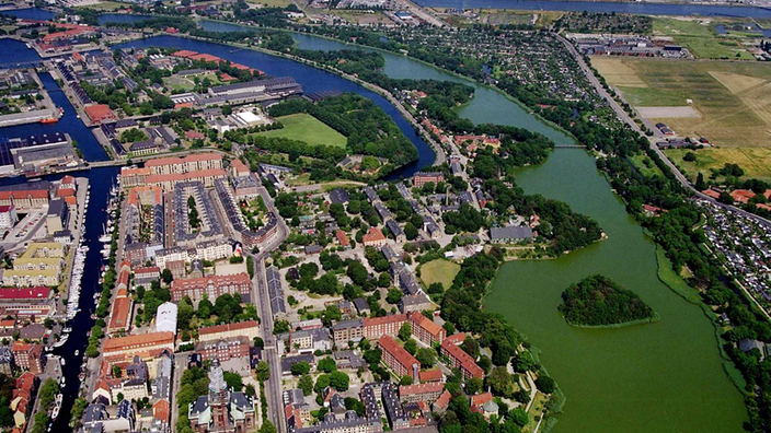 Die Vogelperspektive zeigt den Stadtteil Christiania von Wasser umgeben. Die dichte Bebauung wird durch kleine Grünflächen unterbrochen.