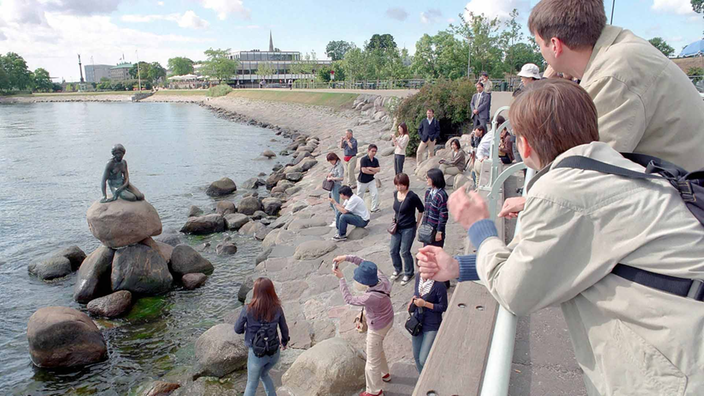 Auf den Natursteinen, die die Uferpromenade zum Meer hin schützen, steigen Touristen herum, um die Skulptur der Meerjungfrau auf dem Steinsockel im Wasser zu fotografieren.
