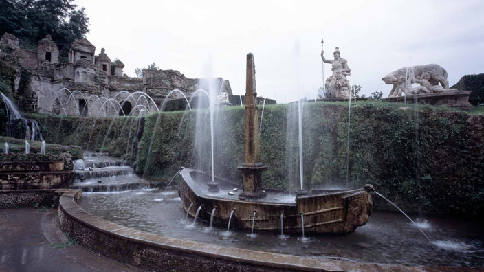 Über vier Stufen fließt Wasser in ein Becken, in dem ein steinernes Schiff als Fontäne dient. Im Hintergrund eine Hecke, über die zahlreiche Fontänen aus dahinterliegenden Wasserspielen spritzen.