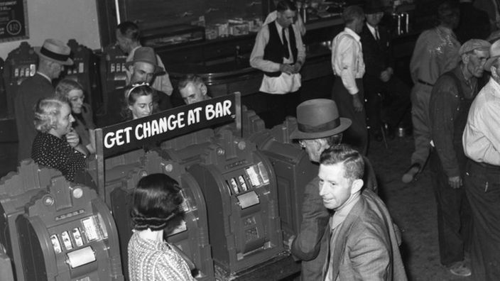 Ein Schwarzweiß-Foto zeigt eine Automatenhalle in Las Vegas im Jahr 1940. Von einem erhöhten Standpunkt aus fotografiert, sieht man Frauen und Männer, die an den Geräten ihr Glück versuchen.