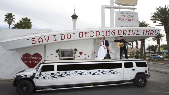 Eine weiße Stretch-Limousine hält vor einem Fenster, über dem 'Wedding Drive Thru' steht