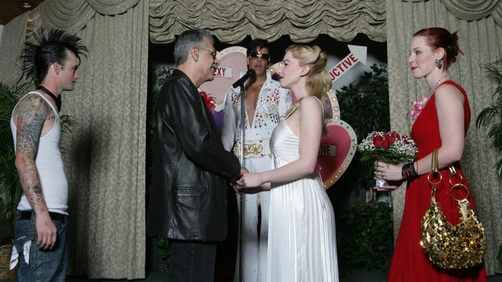 Ein Mann mit Lederjacke und eine Frau im Marilyn-Monroe-Kleid stehen vor einem Elvis-Imitator, links und rechts die Trauzeugen