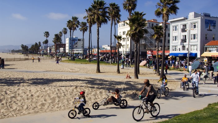 Palmen stehen am Strand von Venice Beach und auf der Promenade fahren viele Bewohner Fahrrad.