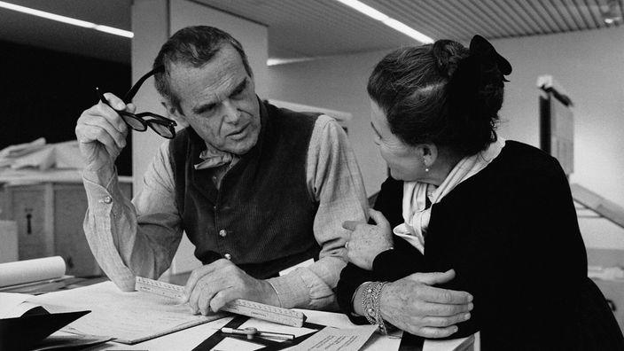 Die Architekten und Designer Charles und Ray Eames sind bei der gemeinsamen Arbeit über einen Zeichentisch gebeugt.
