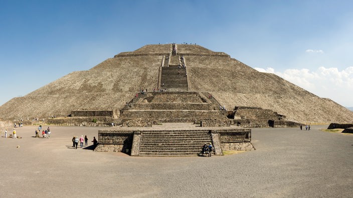 Die mehrstufige Pyramide ist ganz aus grauem Stein erbaut. Wenige Menschen laufen den Weg zu ihr entlang