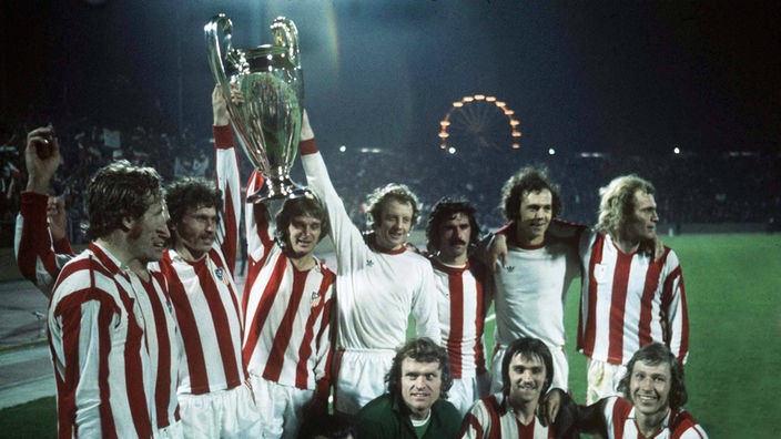 Mannschaftsfoto nach dem Europapokaltriumph von 1974. Die Spieler stehen nebeneinander, drei halten den riesigen Pokal in die Luft.
