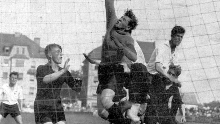 Spielszene von 1928 in Schwarzweiß: Mehrere Spieler in schwarzen und weißen Trikots stehen vor einem Tor. Der Torwart boxt den Ball gerade über die Latte. | Bildquelle: dpa Spielszene von 1928 in Schwarzweiß: Mehrere Spieler in schwarzen und weißen Trikots stehen vor einem Tor. Der Torwart boxt den Ball gerade über die Latte.