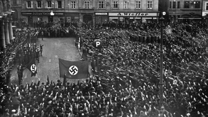 Das Foto zeigt eine Menschenmenge vor einer Bühne, auf der eine Kundgebung stattfindet. Sämtliche Zuschauer halten ihren rechten Arm zum Hitlergruß gehoben. Einige Fahnen mit dem für die Nationalsozialisten typischen Hakenkreuz sind zu sehen.