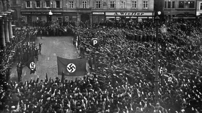 Das Foto zeigt eine Menschenmenge vor einer Bühne, auf der eine Kundgebung stattfindet. Sämtliche Zuschauer halten ihren rechten Arm zum Hitlergruß gehoben. Einige Fahnen mit dem für die Nationalsozialisten typischen Hakenkreuz sind zu sehen. | Bildquelle: akg Das Foto zeigt eine Menschenmenge vor einer Bühne, auf der eine Kundgebung stattfindet. Sämtliche Zuschauer halten ihren rechten Arm zum Hitlergruß gehoben. Einige Fahnen mit dem für die Nationalsozialisten typischen Hakenkreuz sind zu sehen.