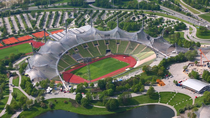 Auf dem Foto ist das Münchener Olympiastadion aus der Vogelperspektive abgebildet. Die Glasdächer über den Zuschauertribünen glänzen in der Sonne. Die Menschen sind nur als Punkte zu erkennen. Im Hintergrund sieht man die Innenstadt Münchens.