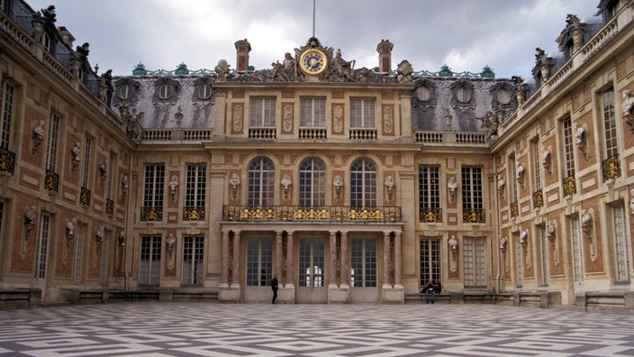 Vor einem gepflasterten Platz mit quadratischen Mustern liegt der Osteingang des Schlosses, zu sehen auch die beiden Seitenflügel.