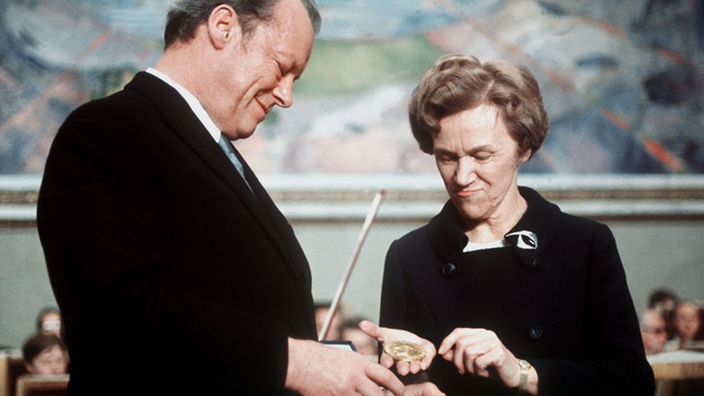 Willy Brandt lächelt stolz, als ihm die schwarz gekleidete Vorsitzende des Nobelpreis-Komitees, Aase Lionaes, den Friedensnobelpreis überreicht | Bildquelle: dpa Willy Brandt lächelt stolz, als ihm die schwarz gekleidete Vorsitzende des Nobelpreis-Komitees, Aase Lionaes, den Friedensnobelpreis überreicht