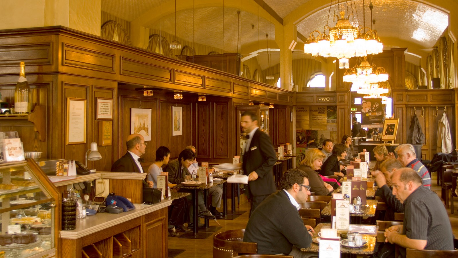 Innenraum des Wiener Café Schwarzenberg. Ein großer Raum mit hoher Decke. An den Tischen sitzen Gäste, die von schwarz livrierten Obern bedient werden.