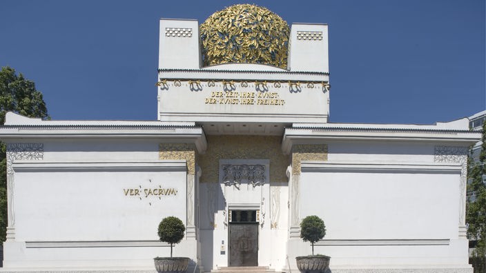 Weißes Gebäude mit goldener Blätterkuppel und Goldverzierungen; über dem Eingang eine Inschrift.