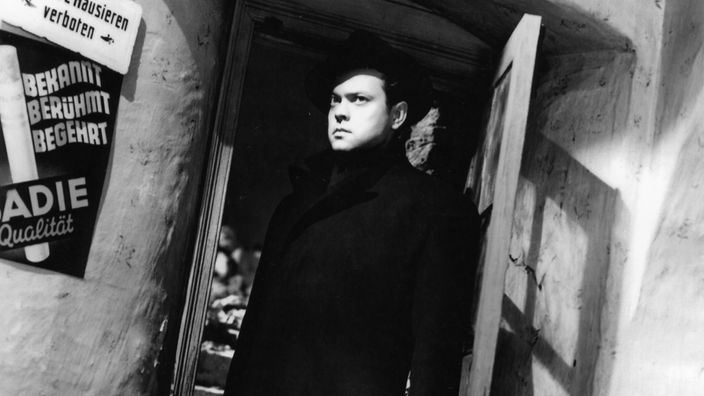 Orson Welles tritt im schwarzen Mantel mit Hut aus einem im Schatten gelegenen Kneipeneingang.