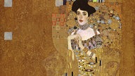 Gemälde "Adele Bloch-Bauer I" von Gustav Klimt: Eine verführerische dunkelhaarige Frau mit einem reich verzierten Kleid vor goldenem Hintergrund.