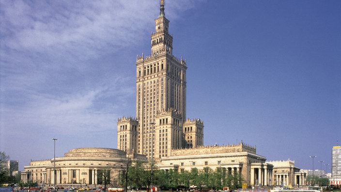 Ein monumentales Gebäude aus dunkelgrauem Stein streckt seinen spitzen Turm in den Himmel, davor ein runder Anbau