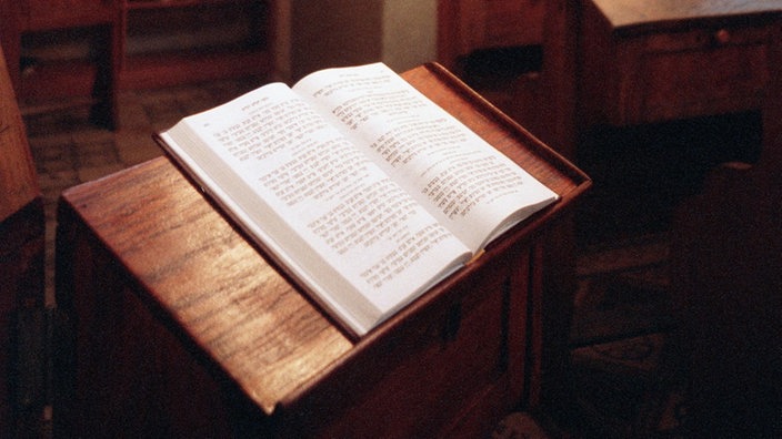 Blick in einen Raum: Auf einem Lesepult liegt im Vordergrund ein aufgeschlagenes Buch, dahinter Bänke, Bücherregale und ein Thora-Schrein.