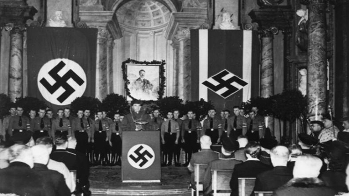 Die Schwarzweiß-Fotografie zeigt den Hörsaal einer Universität bei einer feierlichen Rede. Im Hintergrund ist ein Hitler-Porträt zu sehen, an mehreren Stellen sind Fahnen mit Hakenkreuzen gehisst.
