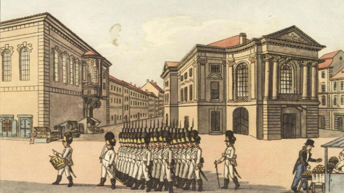 Die kolorierte Radierung zeigt links das Universitätsgebäude mit Erker, rechts das Ständetheater mit Säulen und Bogenfenstern. Auf dem Platz davor marschieren Soldaten.
