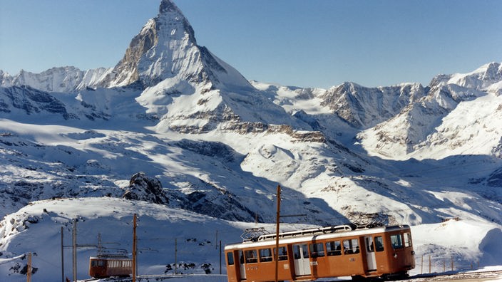Eine rote Zahnradbahn fährt über ein Schneefeld, im Hintergrund das Matterhorn