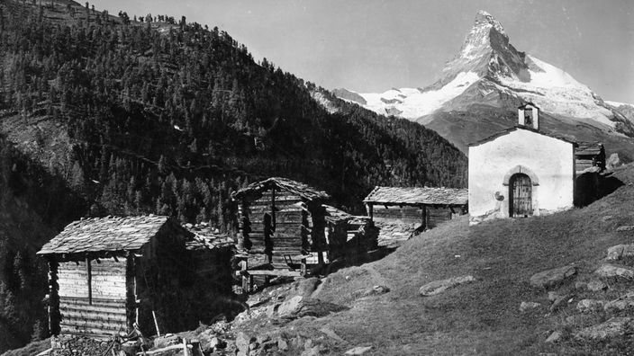 Schwarzweiß-Fotografie eines kleinen Dorfs mit Holzhütten und einer Kapelle direkt unterhalb des Matterhorns